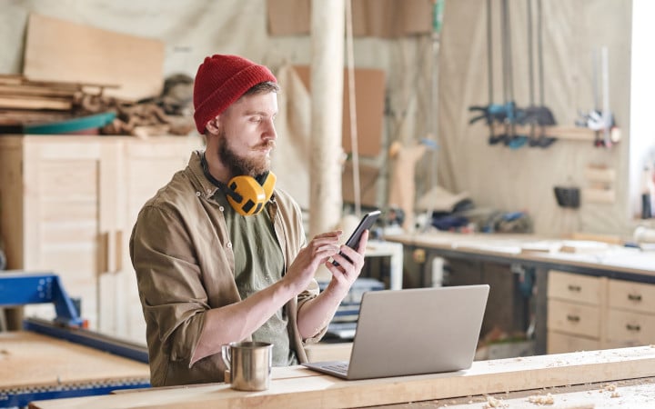 Menuisier moderne debout à son établi avec un ordinateur portable et une tasse de thé envoyant un message sur son smartphone